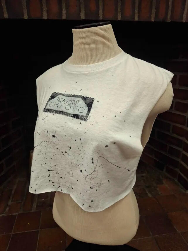 a white tee shirt cut sleeveless and cropped, black and grey paint splattered on the front. It has a small rectangular patch with a grey pattern on the fabric, and the "FEEL CHAOTIC" stamp on it in white. The patch is sewn on with visible cord.