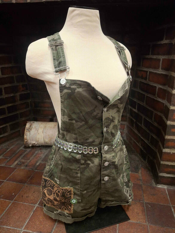Feminine camo overall shorts with soda can tabs stitched on around the waistband and two patches put near the bottom of the right side. One big mushroom + butterfly patch on brown, and one tiny heart patch on green.