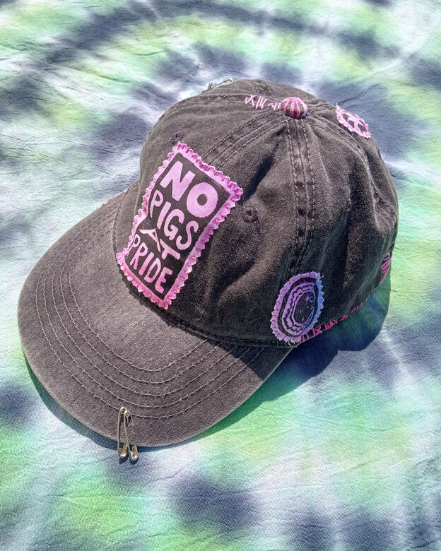 A dark grey baseball style hat on a green black and white tie dye cloth. The hat has pink purple and blue gradient patches of block printed designs stitched on with pink embroidery floss. There are a few small safety pins added as decor, as well as decorat