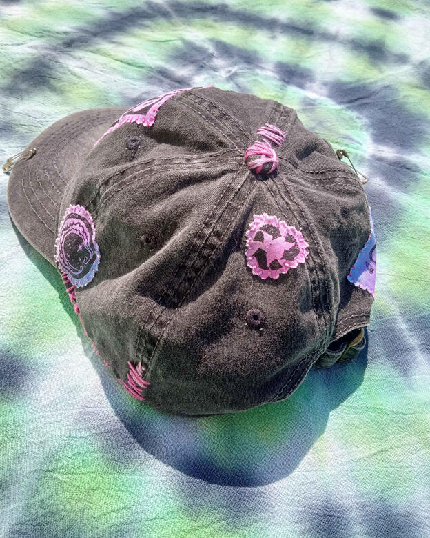 A similar photo of the same hat taken from the back left of it, showing a small pink patch with the recycling symbol, as well as decorative stitching on the top of the hat.